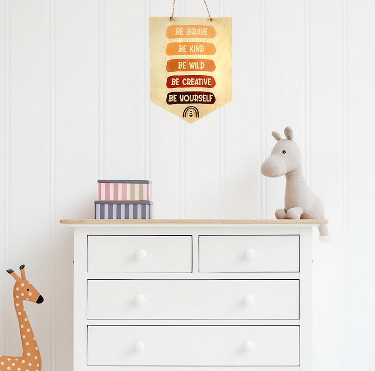 The "Be Brave, Be Kind, Be Creative, Be Wild, Be Yourself" wooden sign hung above a white chest of drawers. Below the sign, a plush horse and two stacked striped boxes are arranged, with a wooden giraffe standing beside the drawers, creating a whimsical and inviting atmosphere.