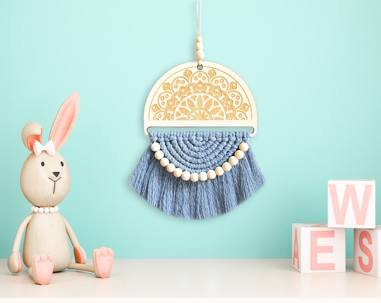 A dusty blue macrame wall hanging displayed on a vibrant aqua wall, positioned between a bunny statue and letter squares, adding a lively contrast.