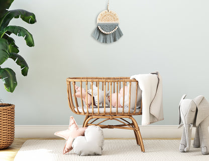 A sage green macrame wall hanging displayed on a light sage wall, placed above a wooden crib. The setting includes a large bird of paradise plant, an oversized elephant toy, and pink and grey cushions, creating a serene and cozy nursery atmosphere.