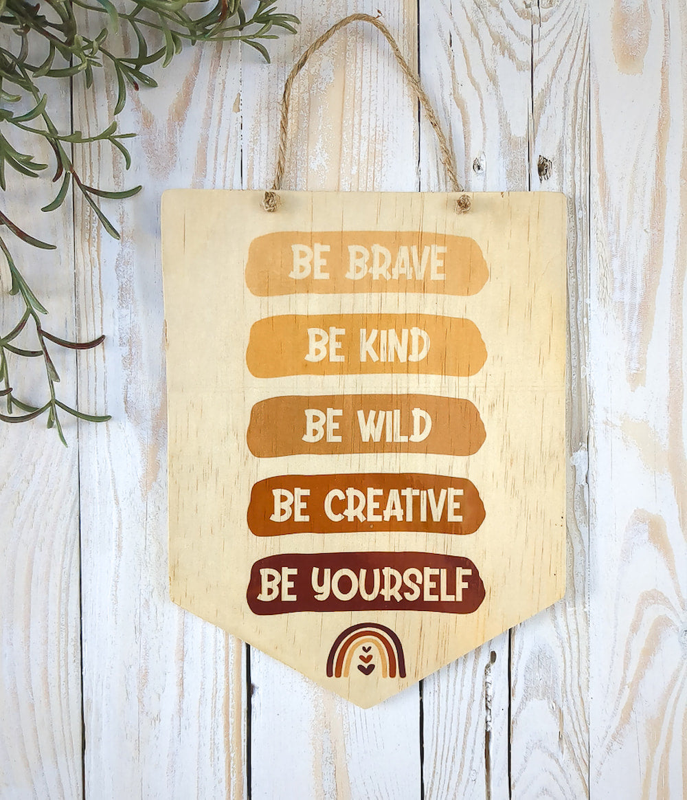 The "Be Brave, Be Kind, Be Creative, Be Wild, Be Yourself" wooden sign resting on white wash timber, surrounded by touches of greenery. 