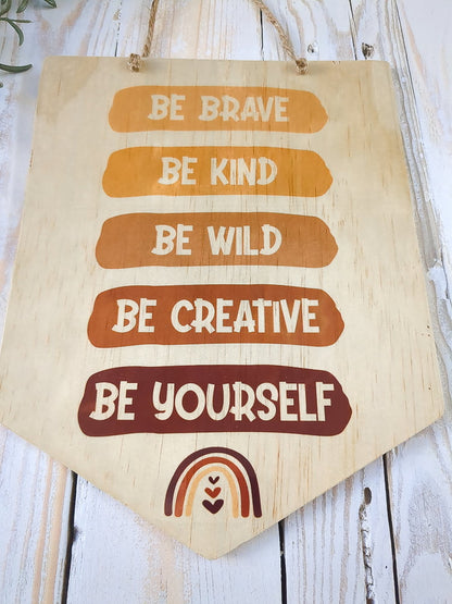 A view from below the "Be Brave, Be Kind, Be Creative, Be Wild, Be Yourself" wooden sign, showcasing its craftsmanship as it rests on white wash timber, with green leaves adding a fresh touch.