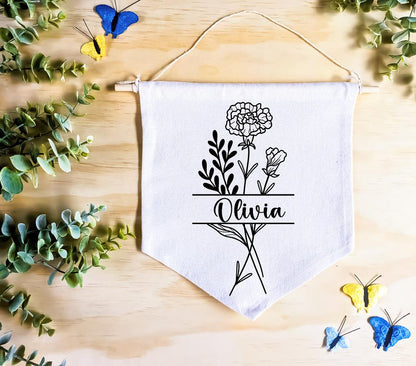 Birth Flower Name Flag made of natural white cotton, featuring the name 'Olivia' with the January birth flower, carnation, both in vivid black. The flag rests on a natural wood surface, surrounded by green leaves and butterfly decor, creating a whimsical, nature-inspired setting.