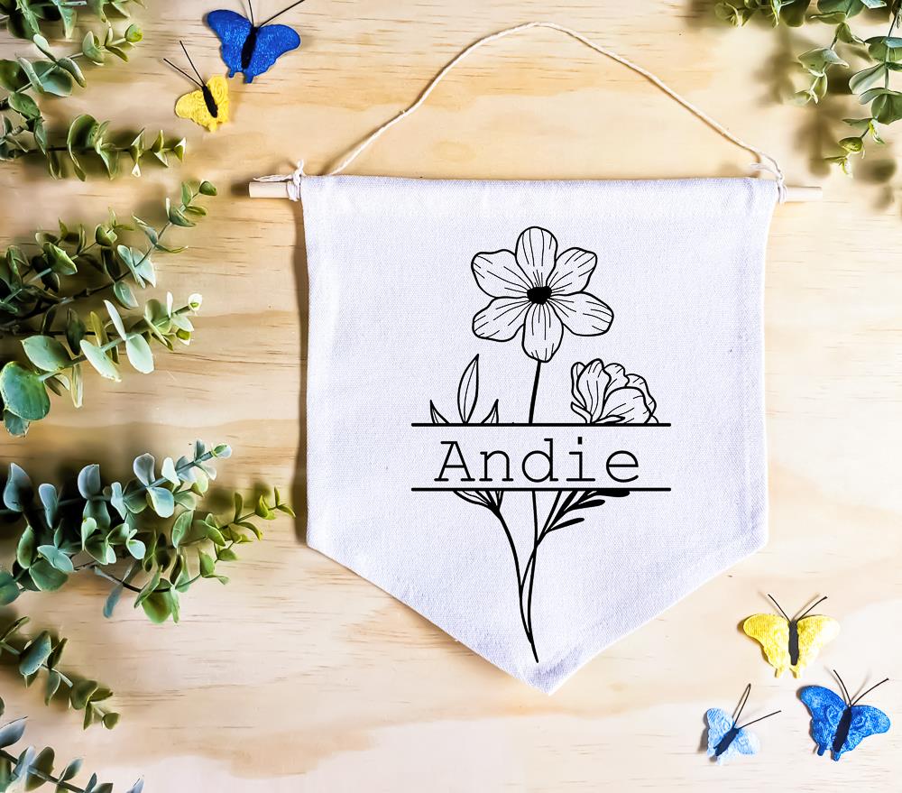 Birth Flower Name Flag made of natural white cotton, featuring the name 'Andie' with the October birth flower, cosmos, both in vivid black. The flag rests on a natural wood surface, surrounded by green leaves and butterfly decor, creating a whimsical, nature-inspired setting.