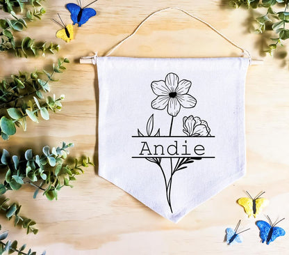 Birth Flower Name Flag made of natural white cotton, featuring the name 'Andie' with the October birth flower, cosmos, both in vivid black. The flag rests on a natural wood surface, surrounded by green leaves and butterfly decor, creating a whimsical, nature-inspired setting.