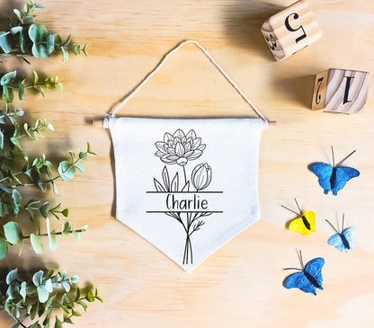 Birth Flower Name Flag made of natural white cotton, featuring the name 'Charlie' with the July birth flower, water lily, both in vivid black. The flag rests on a natural wood surface, surrounded by green leaves and butterfly decor, square wooded numbers, creating a whimsical, nature-inspired setting.