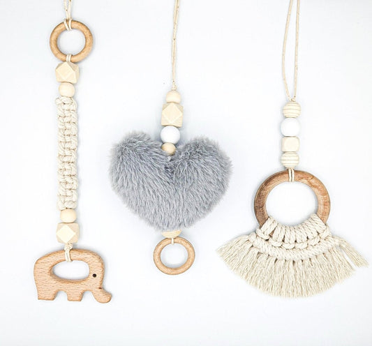 A collection of three Boho macrame play gym toys displayed on a white background: a wooden elephant, a plush grey heart with wooden beads and a ring, and a large wooden circle adorned with macrame tassels and wooden beads.