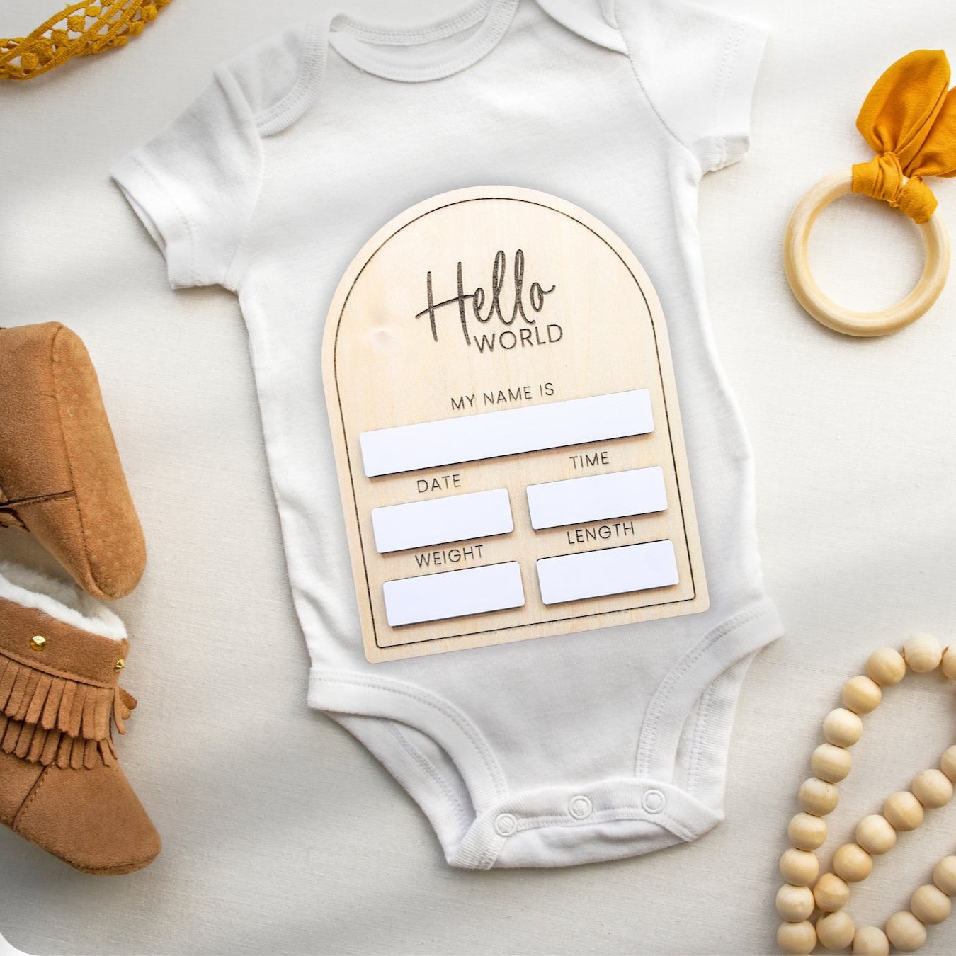 Classic Birth Announcement plaque placed on a baby onesie, accompanied by baby beads, a teether, and baby ugg boots for a cozy, styled setup.