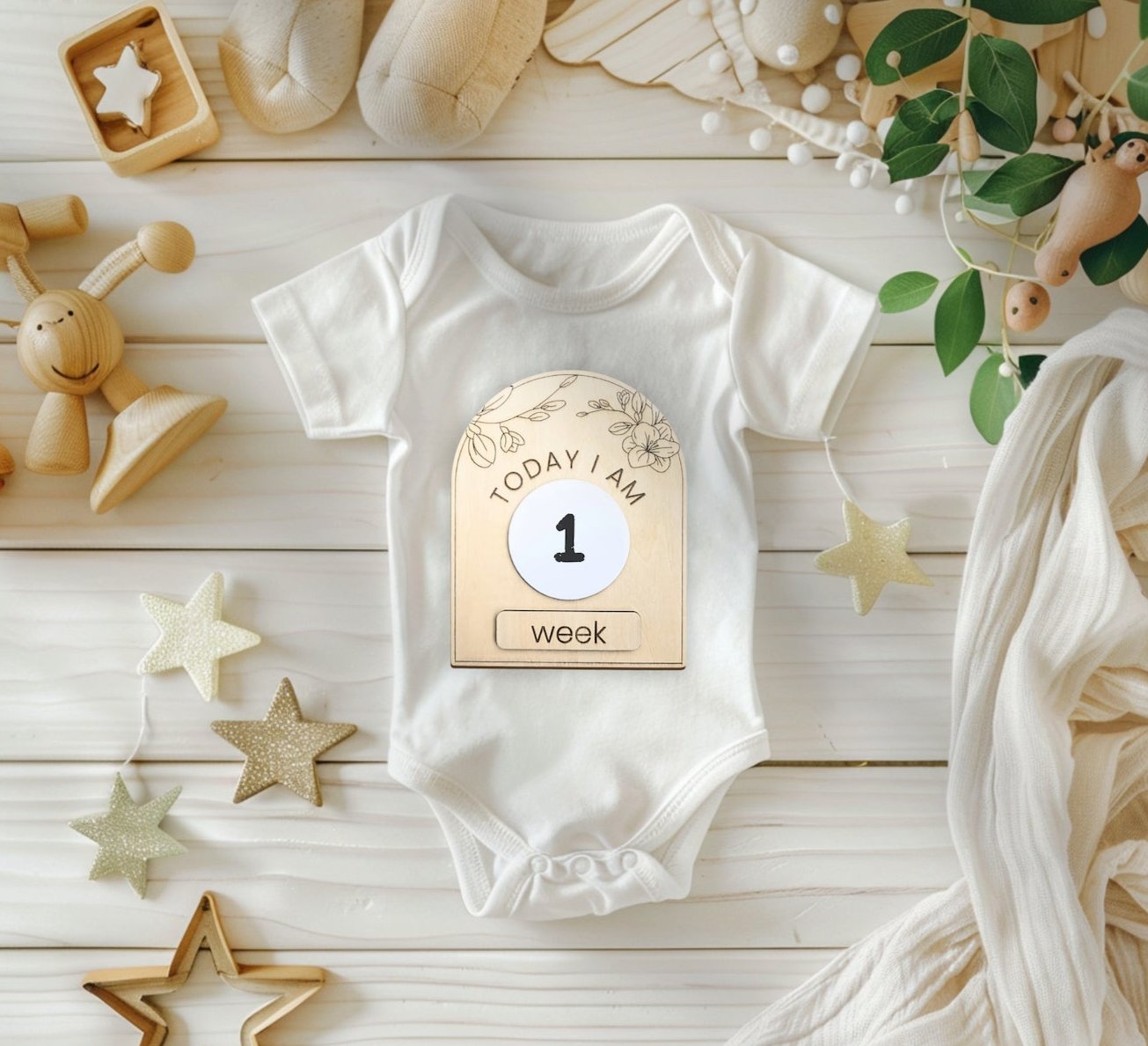 Dry-erase baby milestone plaque reading '1 week,' placed on a baby onesie and surrounded by star cutouts and green foliage for a cozy, natural setting.