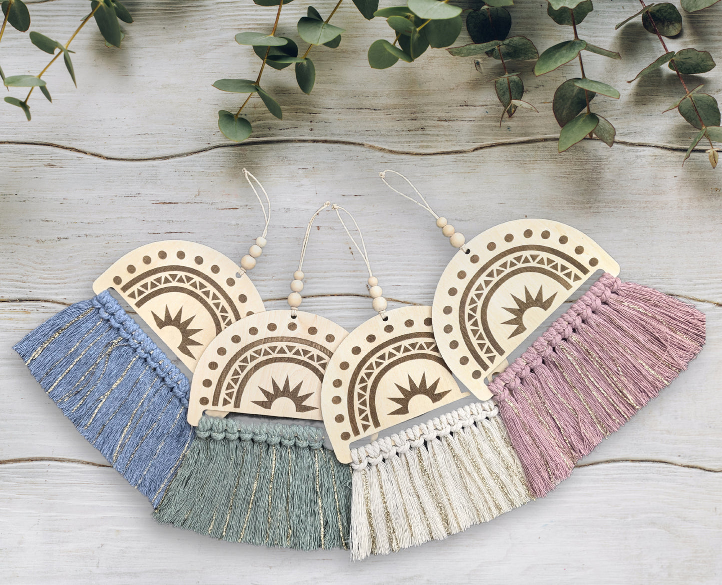 All four Rainbow Macramé Wall Art pieces—natural white, sage green, denim blue, and dusty pink with gold tassels—displayed side by side against a white wooden background with eucalyptus leaves arranged above for a natural, serene setting.