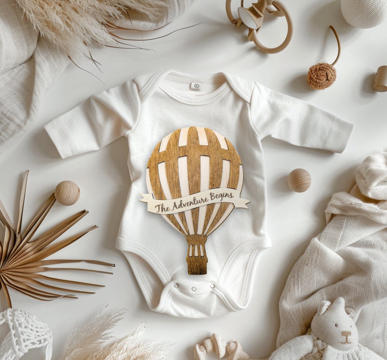 Whimsical Hot Air Balloon Milestone plaque with 'The Adventure Begins' banner, placed on a baby onesie, surrounded by boho elements and cozy blankets for a warm, stylish setting.