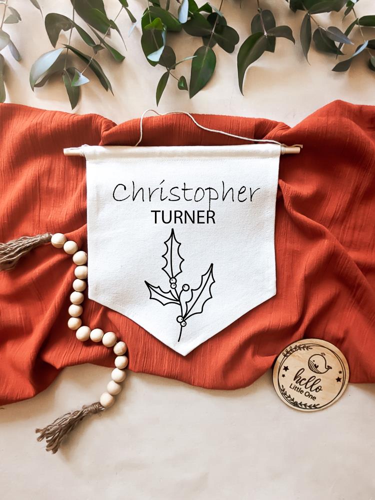 Birth Flower Name Flag made of natural white cotton, displaying the name 'Christopher Turner' with the December birth flower, holly, beneath it. The flag is laid on a burnt red material, with eucalyptus leaves positioned above. Beside the flag are wooden beads and a round wooden disc reading 'Hello Little One".