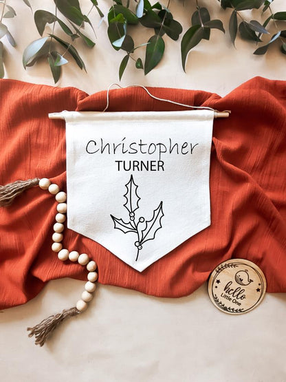 Birth Flower Name Flag made of natural white cotton, displaying the name 'Christopher Turner' with the December birth flower, holly, beneath it. The flag is laid on a burnt red material, with eucalyptus leaves positioned above. Beside the flag are wooden beads and a round wooden disc reading 'Hello Little One".