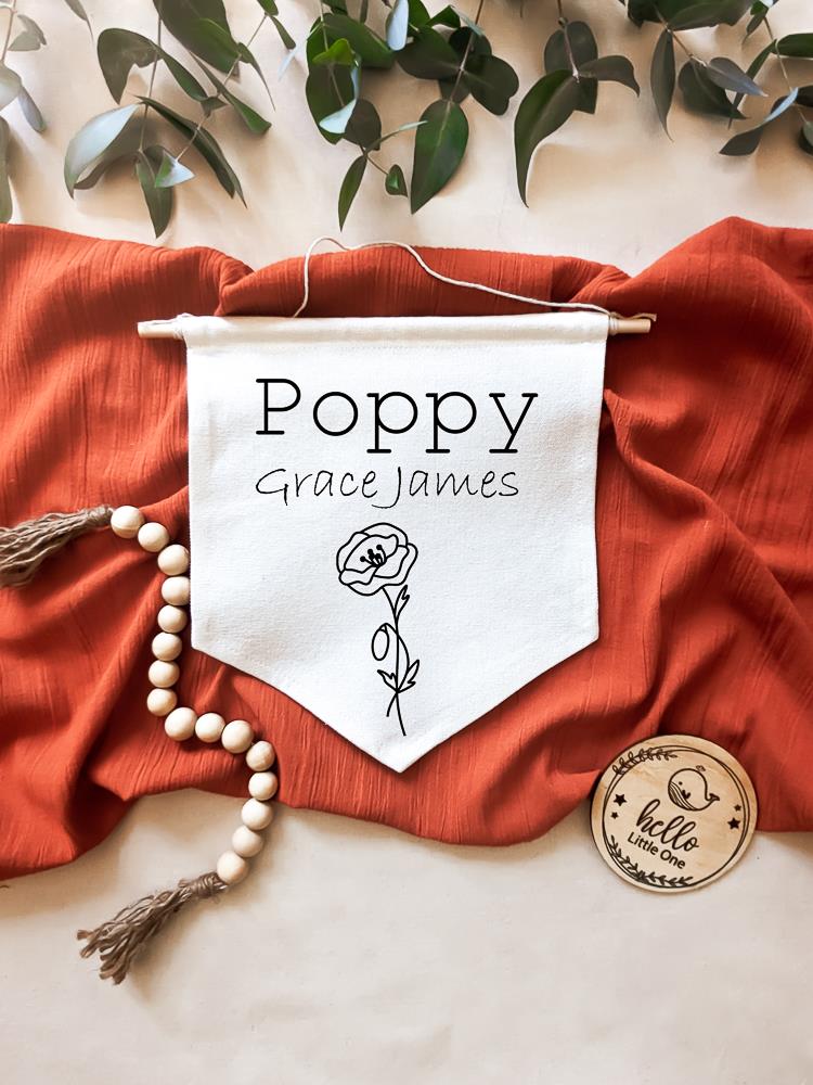 Birth Flower Name Flag made of natural white cotton, showcasing the name 'Poppy Grace James' with the August birth flower, poppy, beneath it. The flag is laid on a burnt red material, with eucalyptus leaves positioned above. Beside the flag are wooden beads and a round wooden disc reading 'Hello Little One.'