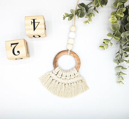  A  wooden circle macrame play gym toy, adorned with macrame tassels, placed next to green leaves and wooden number squares, accentuating its beautiful design and natural aesthetic.