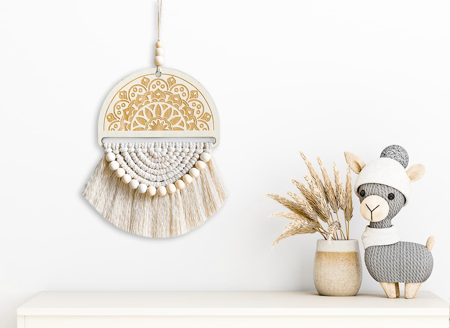 A natural white macrame wall hanging mounted above a shelf, styled with a ceramic sheep statue and dried foliage for a cozy, rustic look.